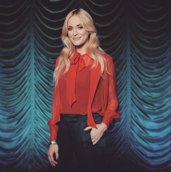 Portrait of a woman with long blonde hair wearing a red blouse and black pants posing in front of a draped turquoise backdrop.