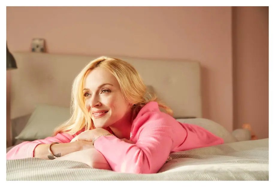 Blonde woman wearing a pink top lying down on a bed in a bedroom setting