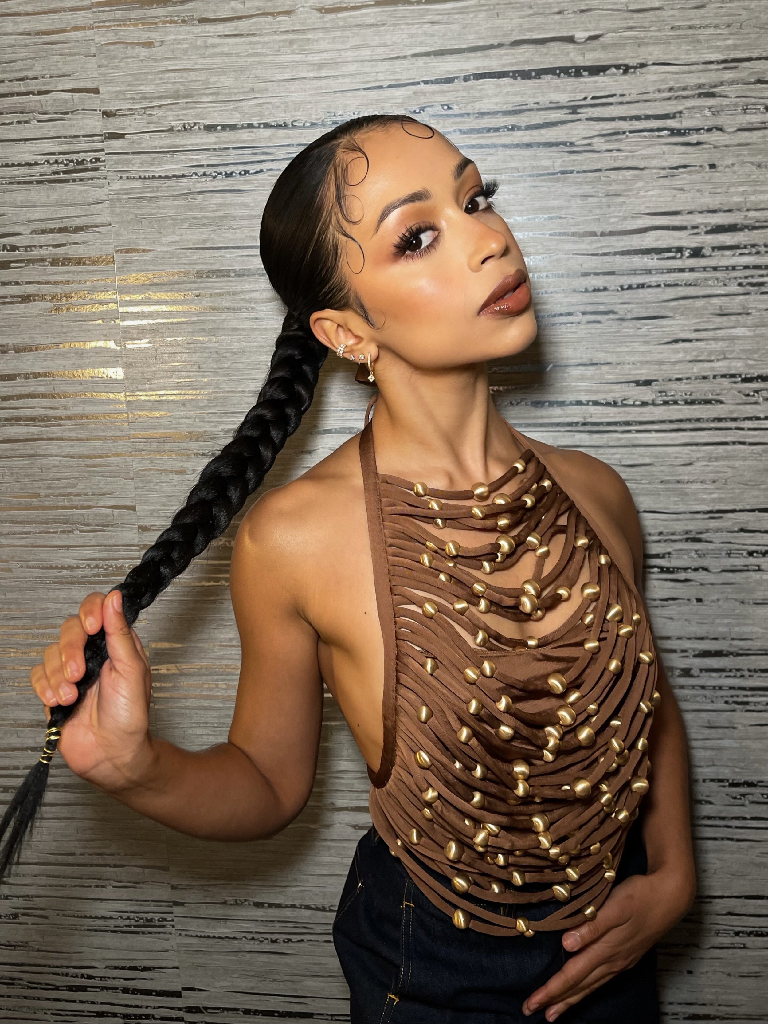 A woman with dark hair in a ponytail holds the end of her braid while posing in a halter top in front of a textured wall.