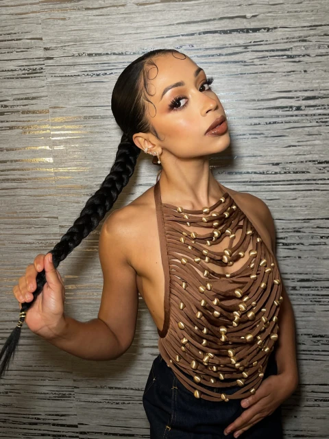 A woman with dark hair in a ponytail holds the end of her braid while posing in a halter top in front of a textured wall.