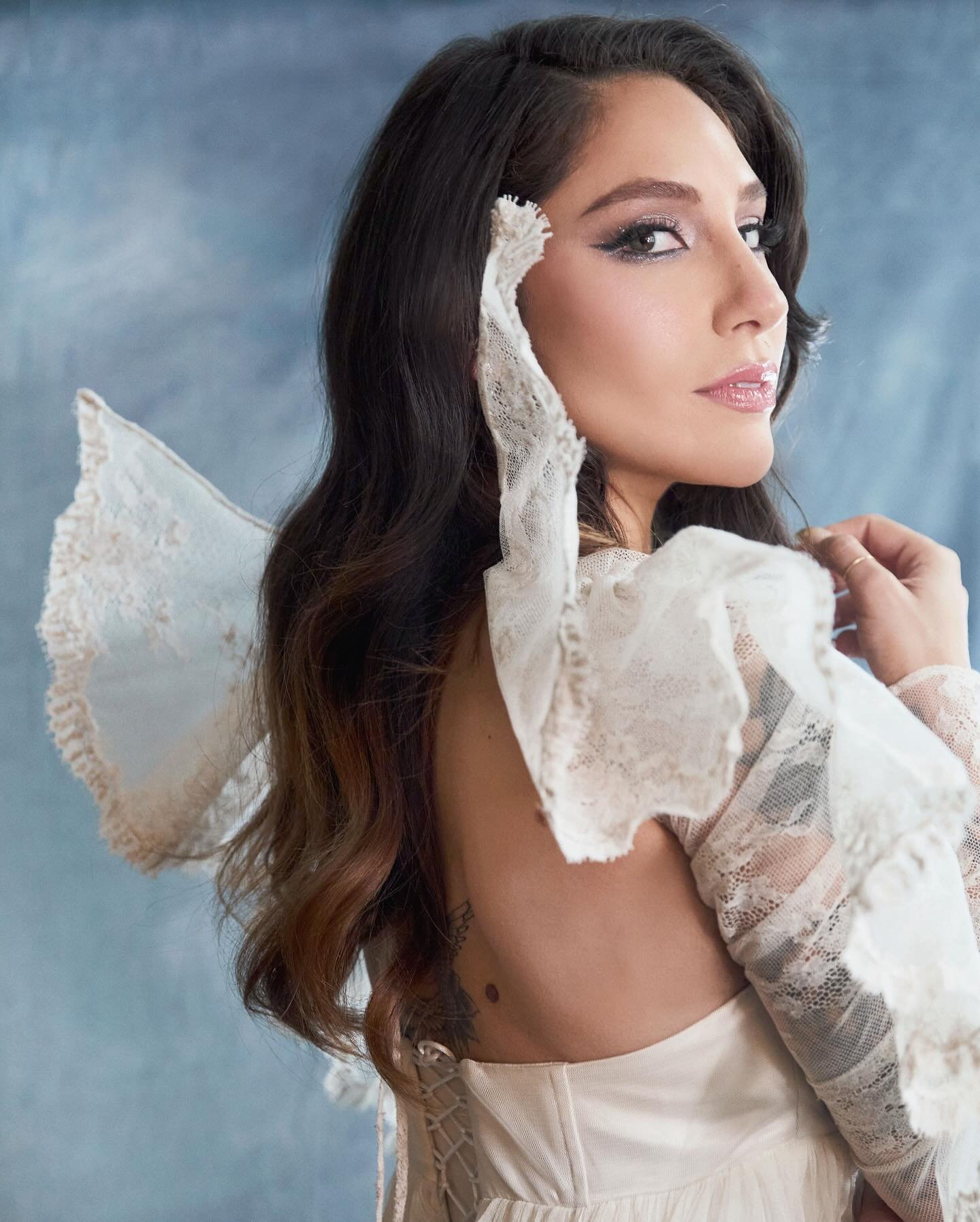 Portrait of a woman with dark hair in an off-white dress with lace sleeves against a blue background in a photo studio setting.