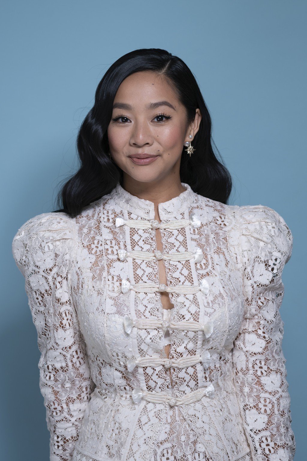 Headshot of an Asian woman with dark wavy hair wearing a white lace dress against a light blue backdrop, captured in a studio setting.