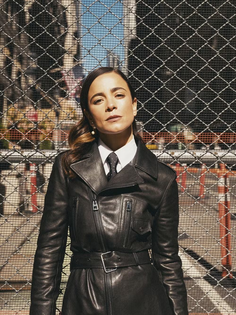 Woman with dark hair, wearing a black leather trench coat, poses in front of a chain link fence with city buildings in the background.