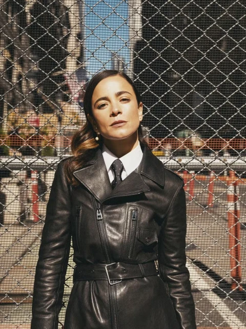Woman with dark hair, wearing a black leather trench coat, poses in front of a chain link fence with city buildings in the background.