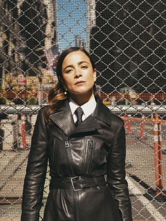 Woman with dark hair, wearing a black leather trench coat, poses in front of a chain link fence with city buildings in the background.