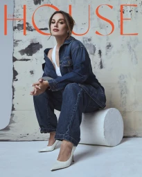 Woman in denim jacket and jeans seated on white cylinder with white heels against distressed wall
