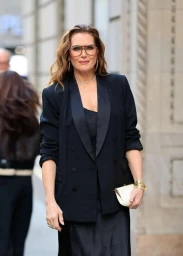 Brooke Shields wearing large tinted glasses, a black blazer, and holding a small white clutch on a city street