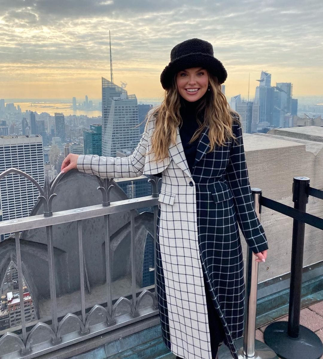 Adult woman in a black and white patterned coat and black hat stands on an observation deck with a cityscape in the background.