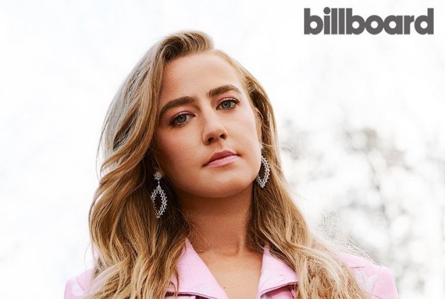 Close-up portrait of a woman with long blonde hair, pink jacket, and jewelry, set against a blurred background. Billboard logo visible.
