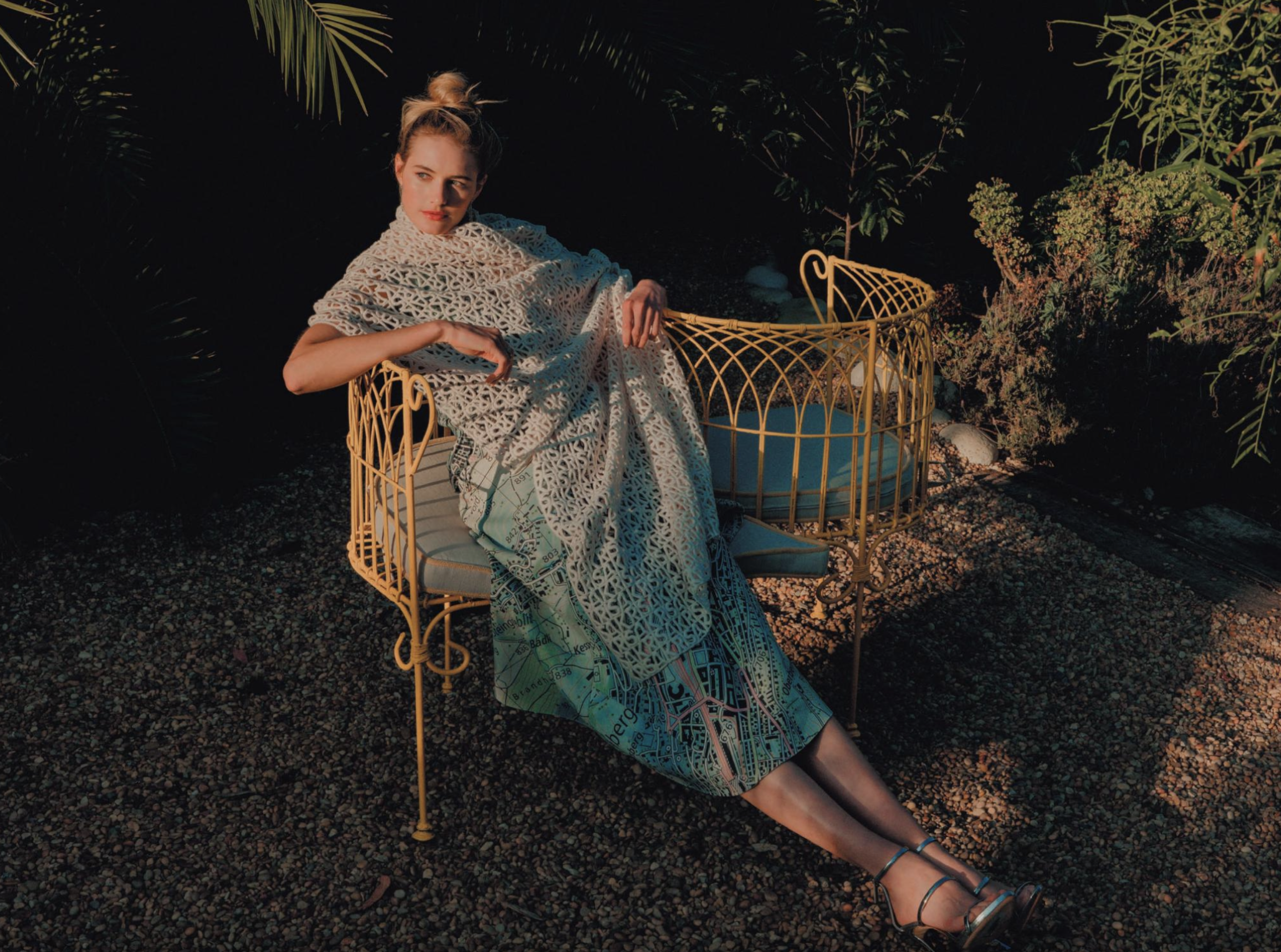 Woman sits on a decorative chair outdoors in a sunlit garden, wearing a lacy wrap and strappy heels in a fashion editorial photo.