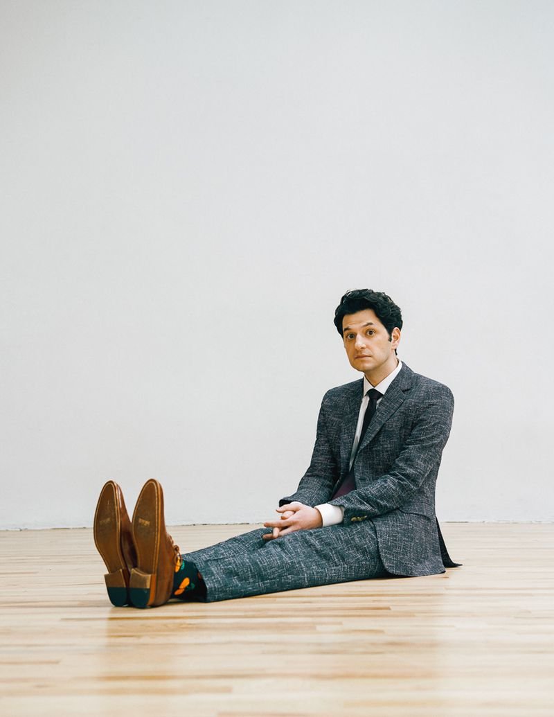 Adult male in a grey suit sits on a wood floor against a white wall, legs extended, hands clasped, wearing colorful patterned socks.