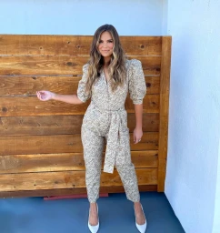 Woman in patterned jumpsuit with puffed sleeves and white heels standing outdoors against wood fencing