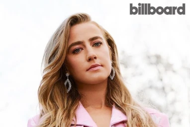 Portrait of singer Lennon Stella wearing a pink patterned jacket and ornate drop earrings against a light background.