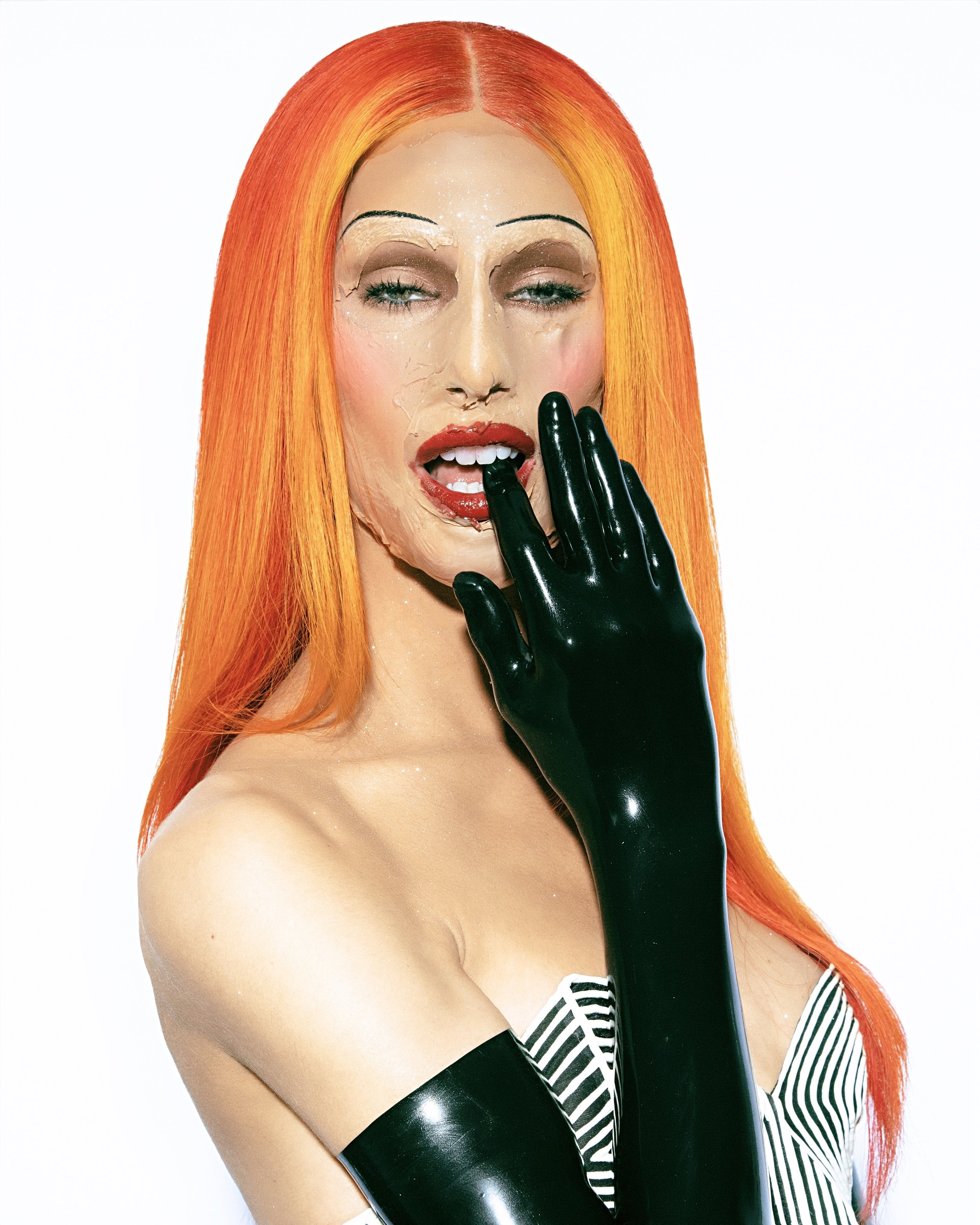 Person with orange hair and theatrical makeup wearing black latex gloves and a black and white striped top in a studio shot.