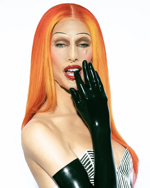 Person with orange hair and theatrical makeup wearing black latex gloves and a black and white striped top in a studio shot.