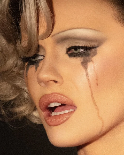 Close-up portrait of a woman with theatrical makeup, including heavy eyeliner smudged to look like tears, against a dark backdrop.