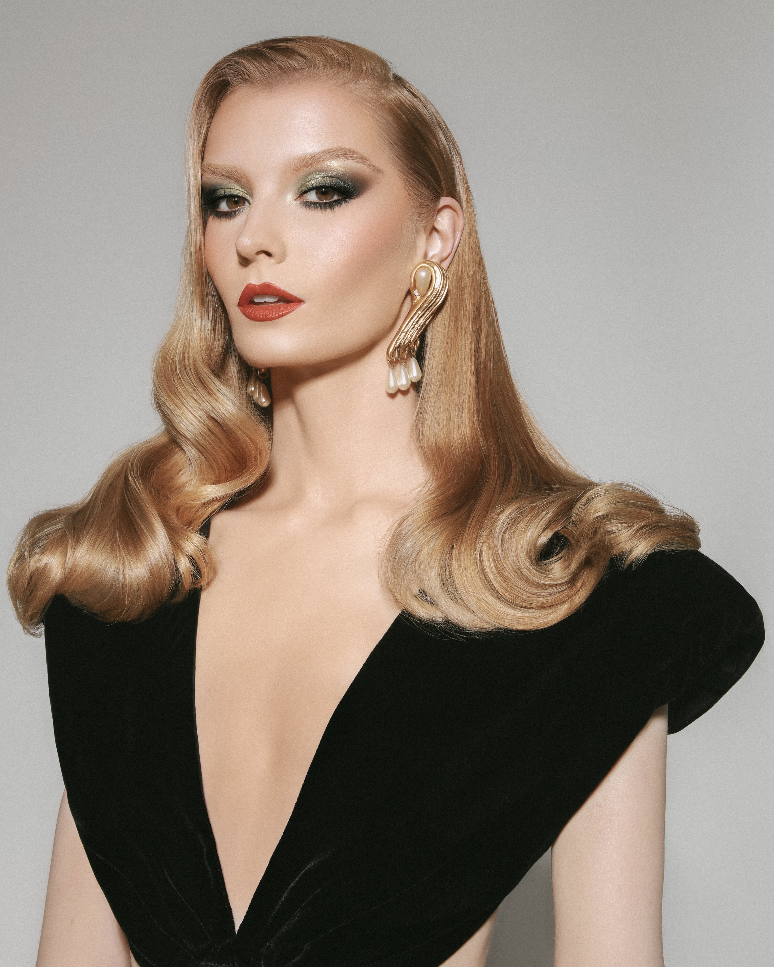 Close-up shot of a blonde woman with glamorous makeup, vintage-styled hair, and pearl earrings wearing a black velvet low-cut dress on gray backdrop.