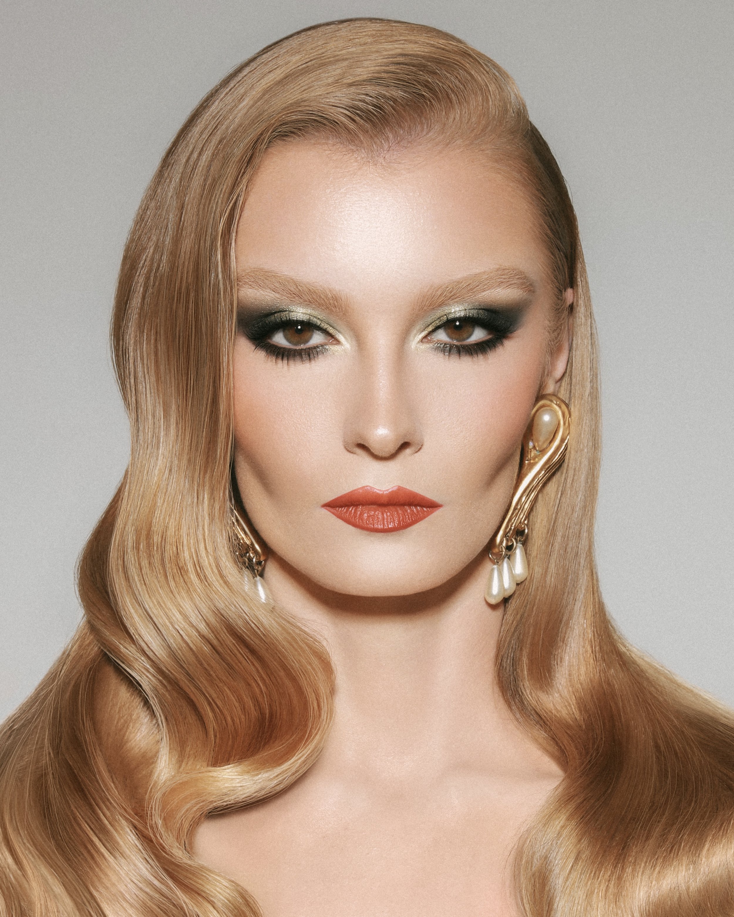 Close-up portrait of a blonde female with striking makeup, including green eyeshadow and orange lipstick, wearing vintage gold earrings.