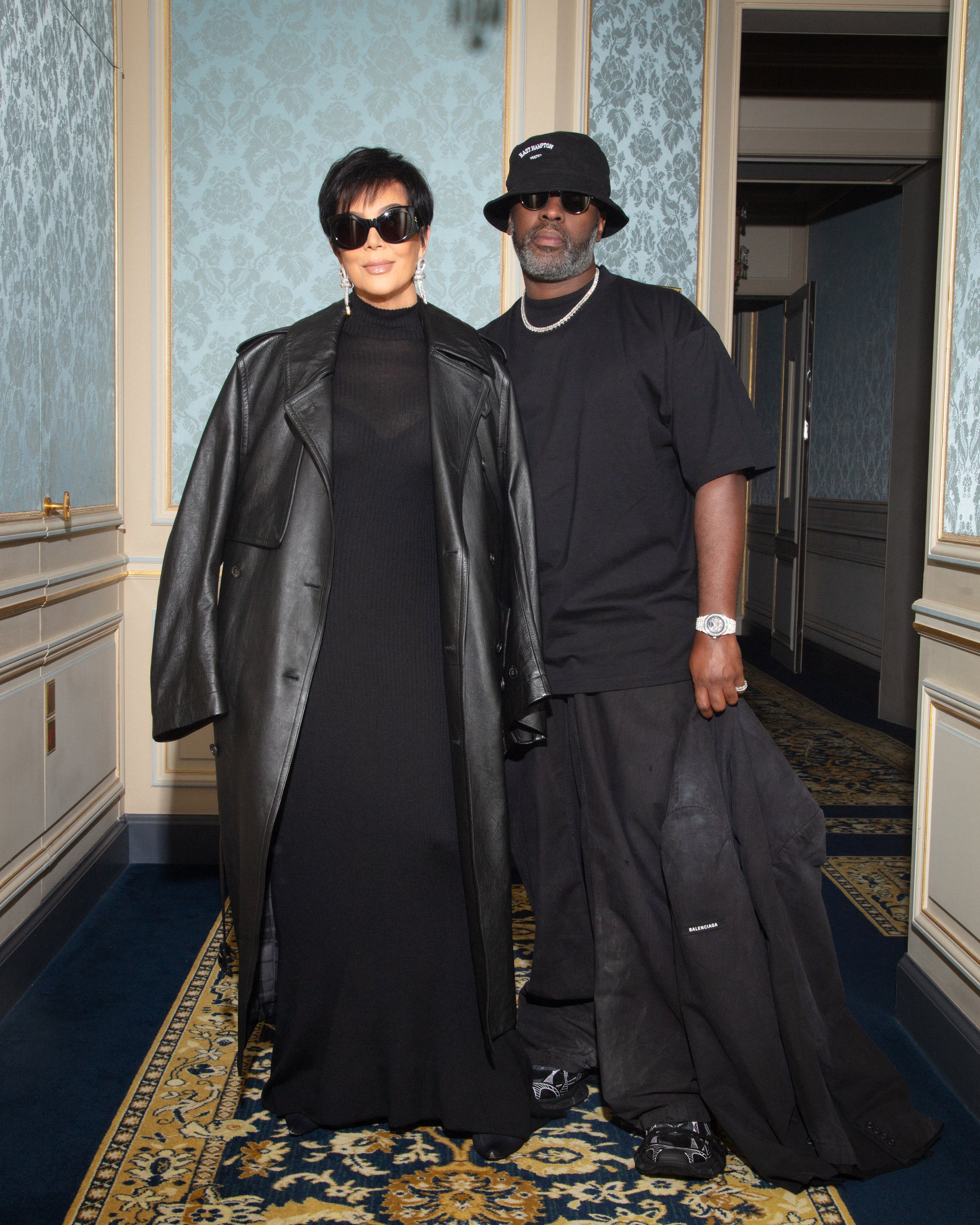 Kris Jenner and Corey Gamble posing indoors against a blue patterned wall, both in dark sunglasses and black clothing.