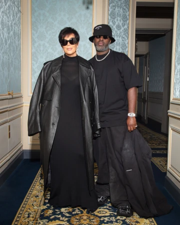 Kris Jenner and Corey Gamble posing indoors against a blue patterned wall, both in dark sunglasses and black clothing.