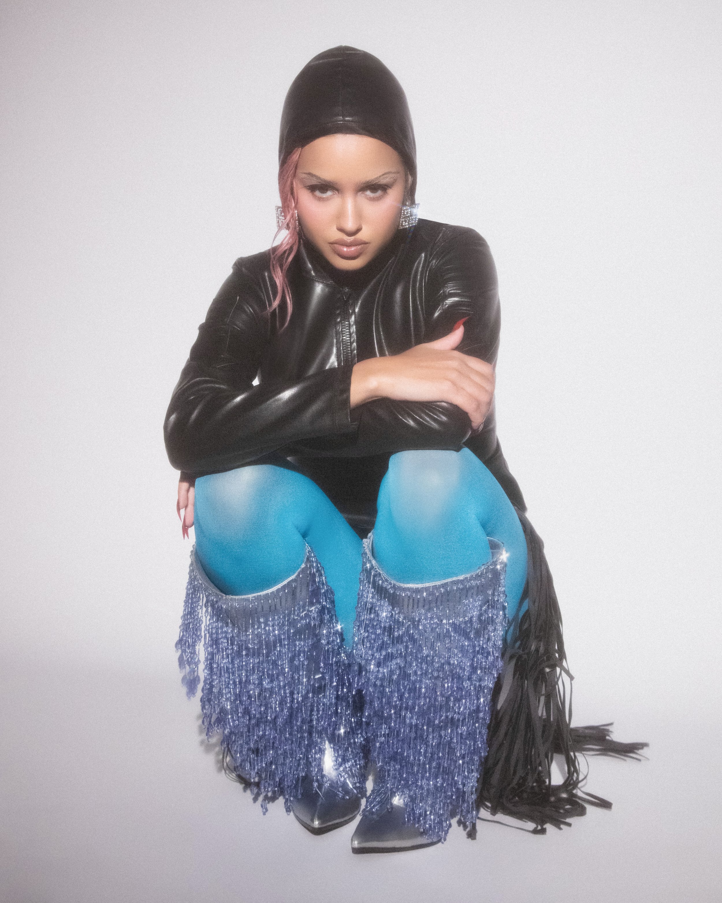 A young adult woman with pink-tinged hair wearing a black leather hooded outfit and turquoise tights with silver fringe boots posing in a studio.