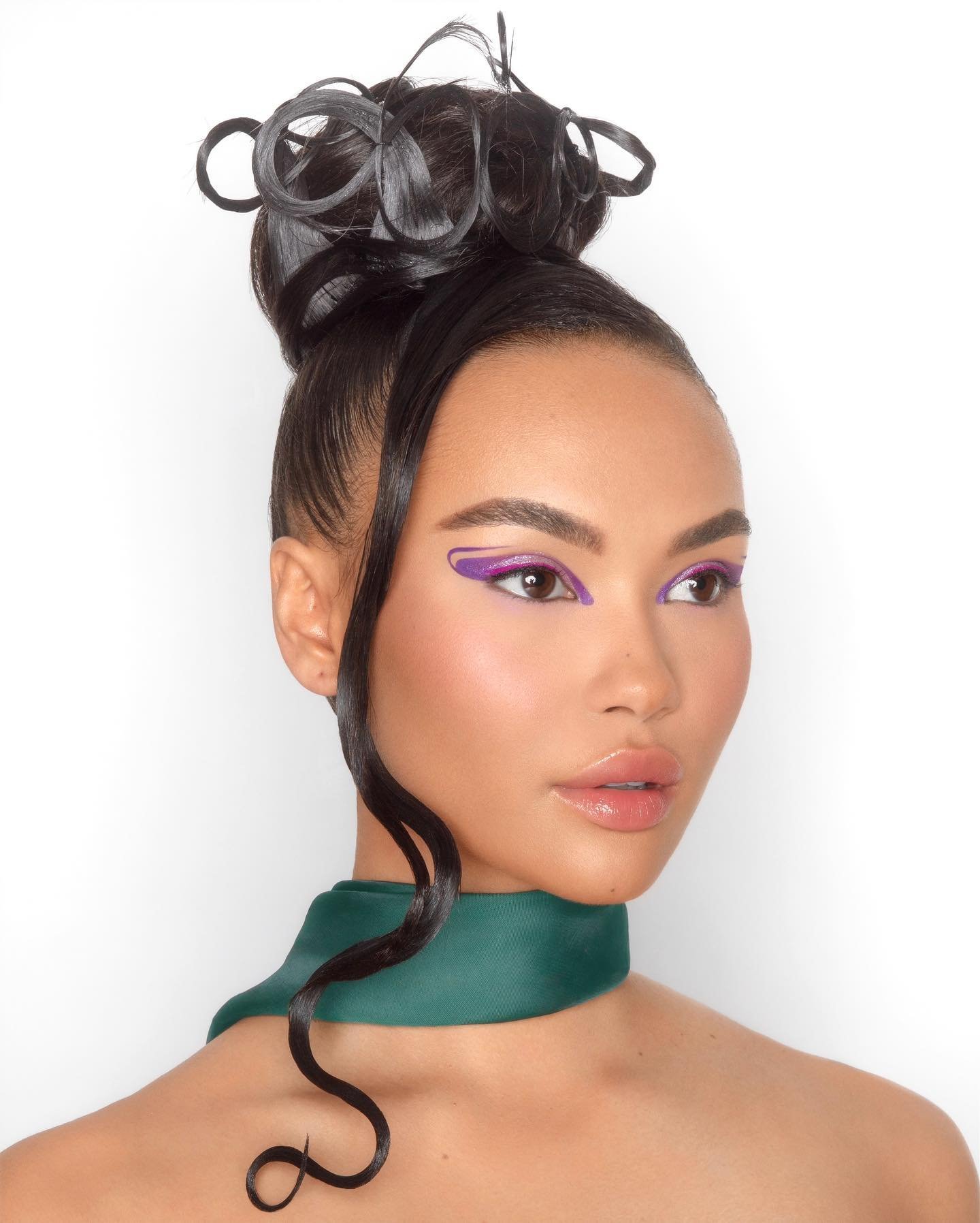 Close-up of a woman with dark hair in a high bun, wearing purple eyeliner and a green choker against a white studio backdrop.