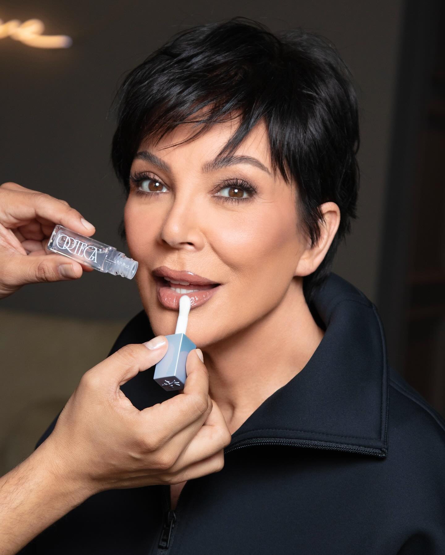 Kris Jenner applying lip gloss in a studio setting with makeup artist visible, beauty shot, cosmetics, skincare promotion.