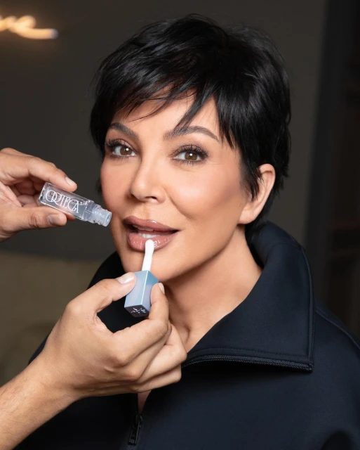 Kris Jenner applying lip gloss in a studio setting with makeup artist visible, beauty shot, cosmetics, skincare promotion.