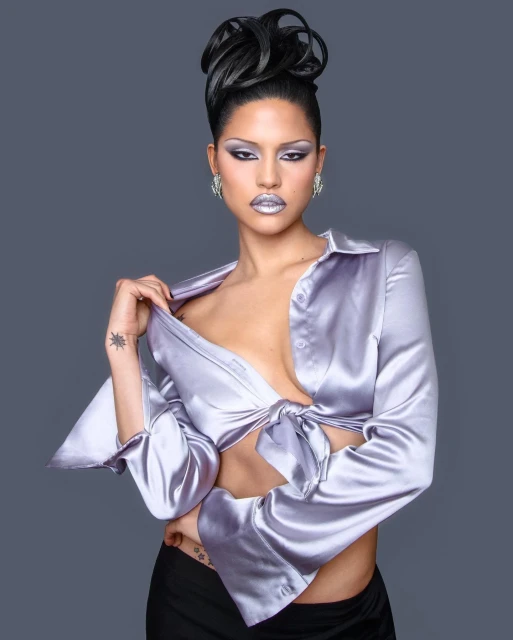 Portrait of a woman with creative hair and makeup, wearing a satin top and dark pants, against a solid grey background.