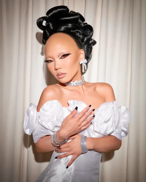 Elegant woman with artistic hair and makeup poses in off-shoulder gown with diamond jewelry against a neutral curtain backdrop in a studio.