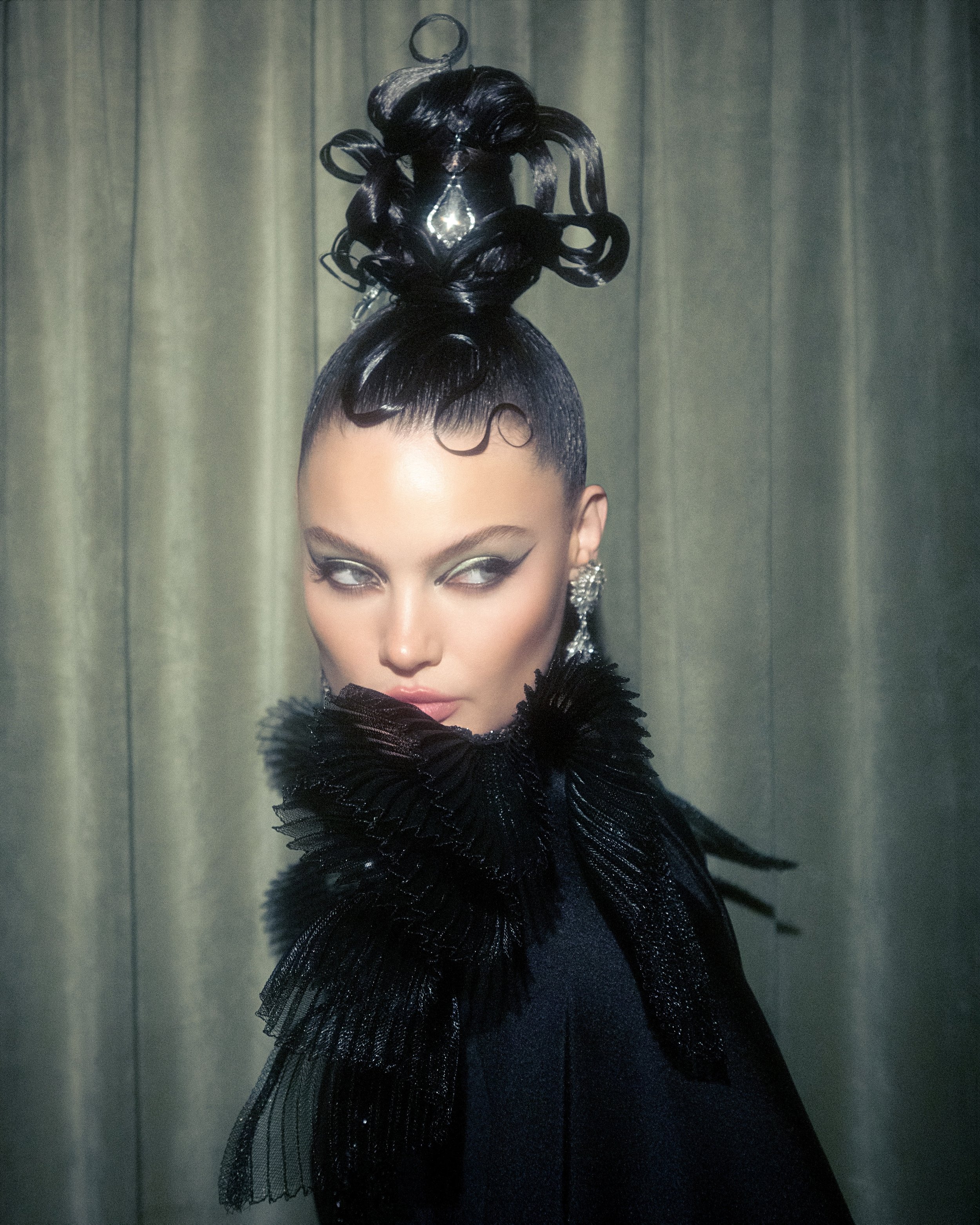Close-up portrait of an adult female with black hair styled up high, wearing statement earrings and a black feathered collar.