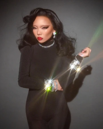 Portrait of a woman wearing a black top and sparkling diamond jewelry against a dark gradient background.