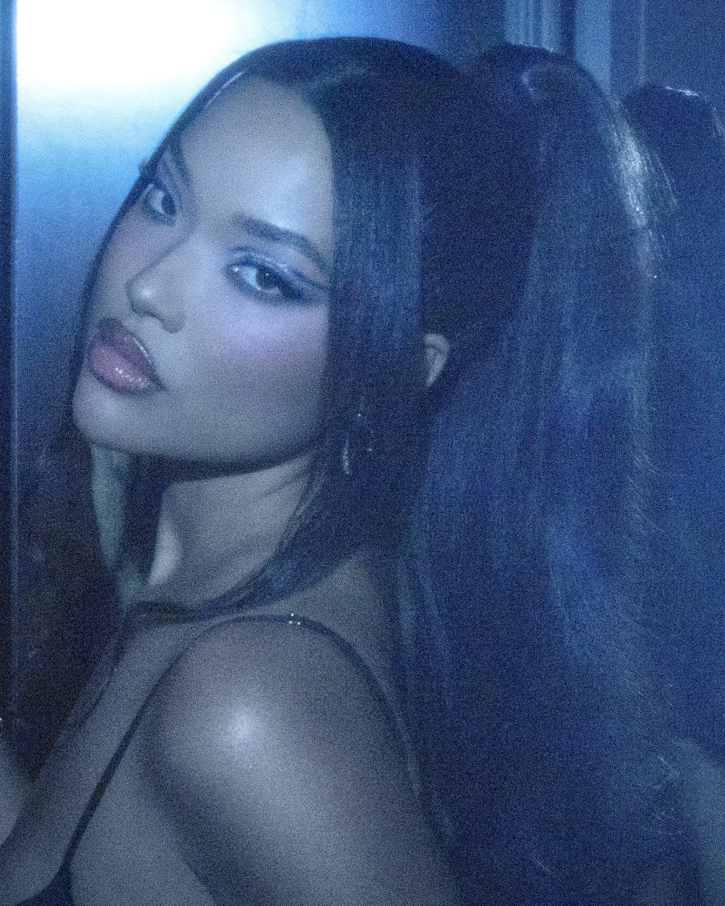 Close-up of a woman with a high ponytail, wearing blue eyeshadow, and posing indoors in front of a blue wall with subtle lighting.