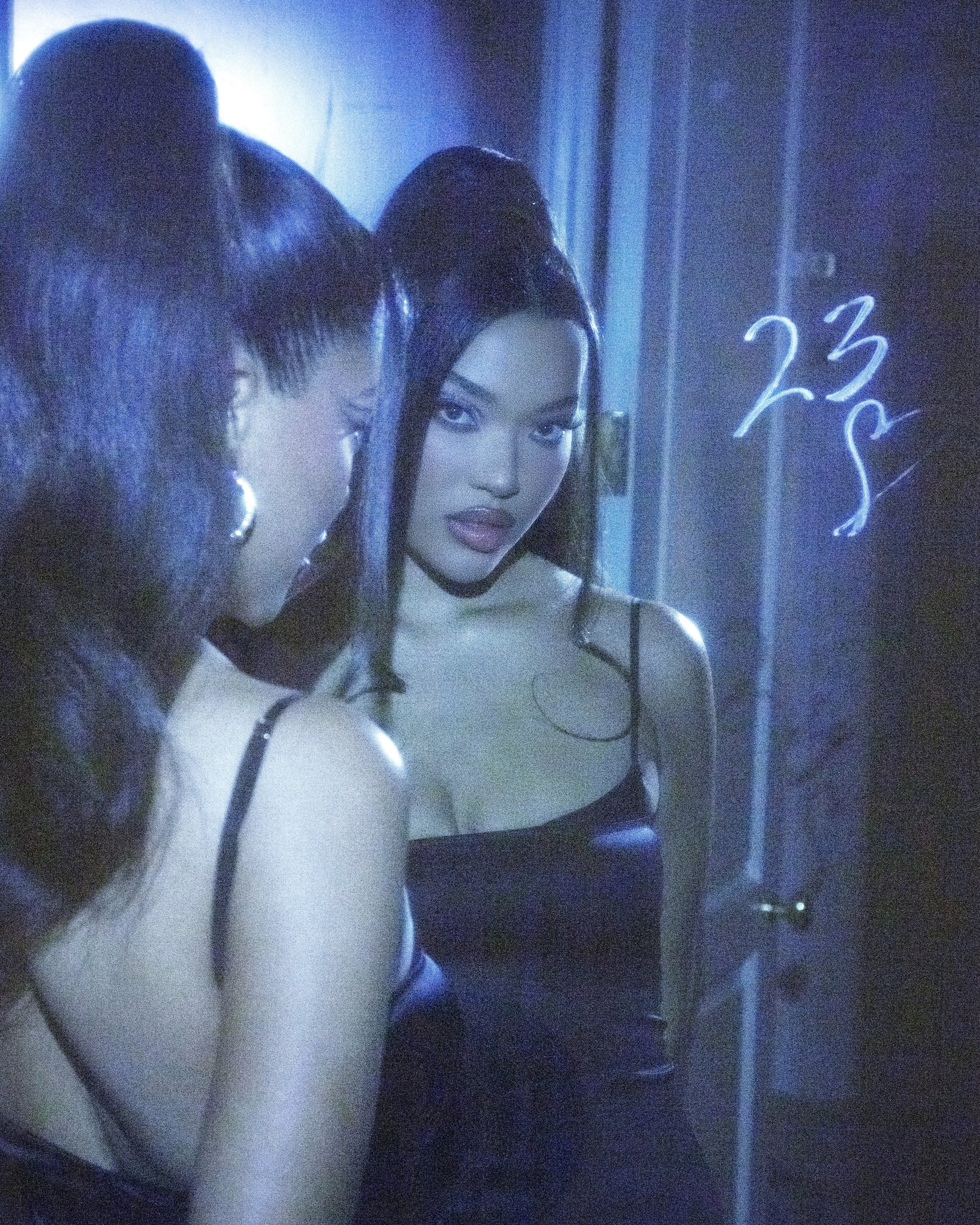 An adult female poses in front of a mirror in a room with moody lighting. She wears a black dress and gold hoop earrings.