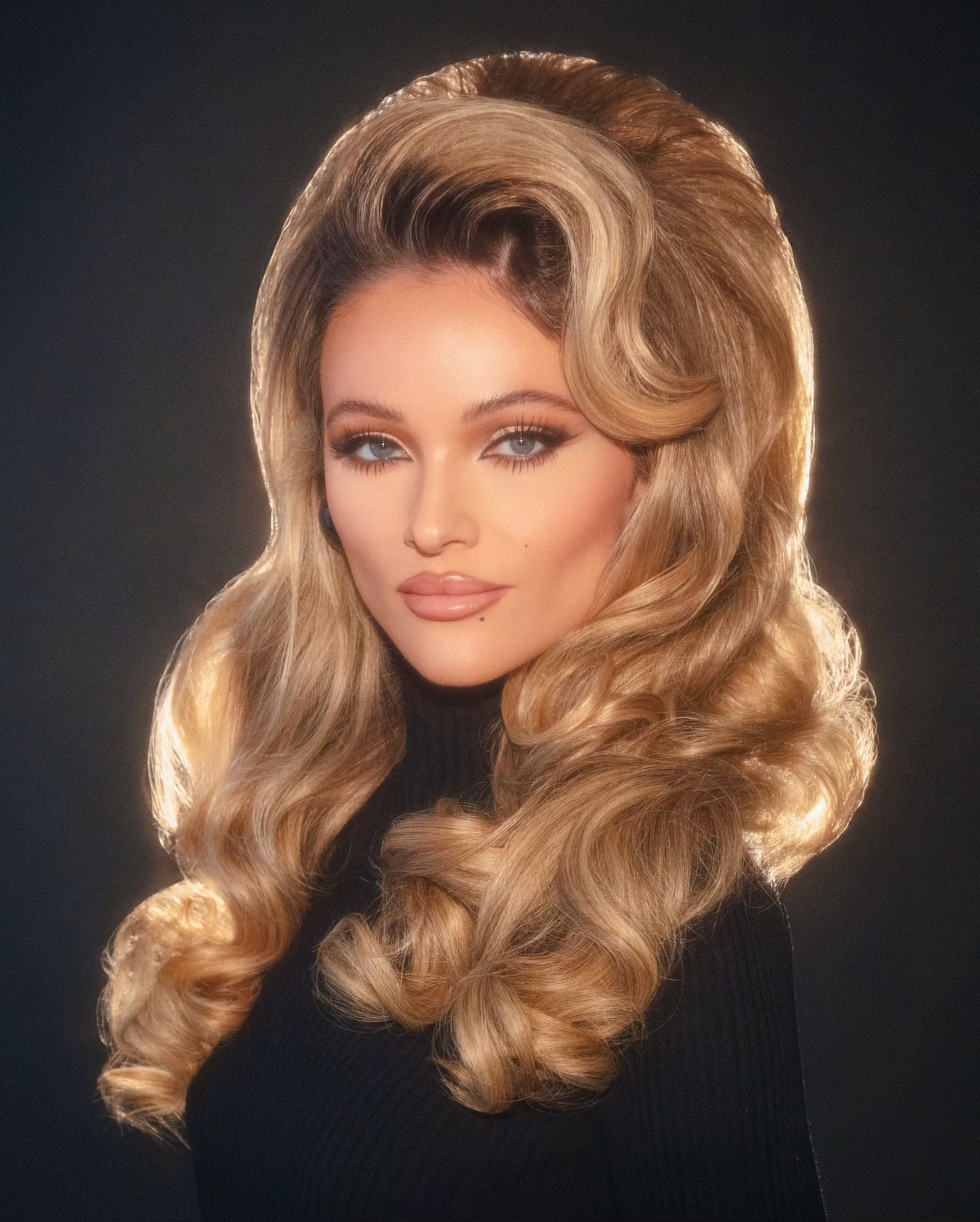 Portrait of a blonde woman with voluminous retro hairstyle and winged eyeliner wearing a black top against a dark background