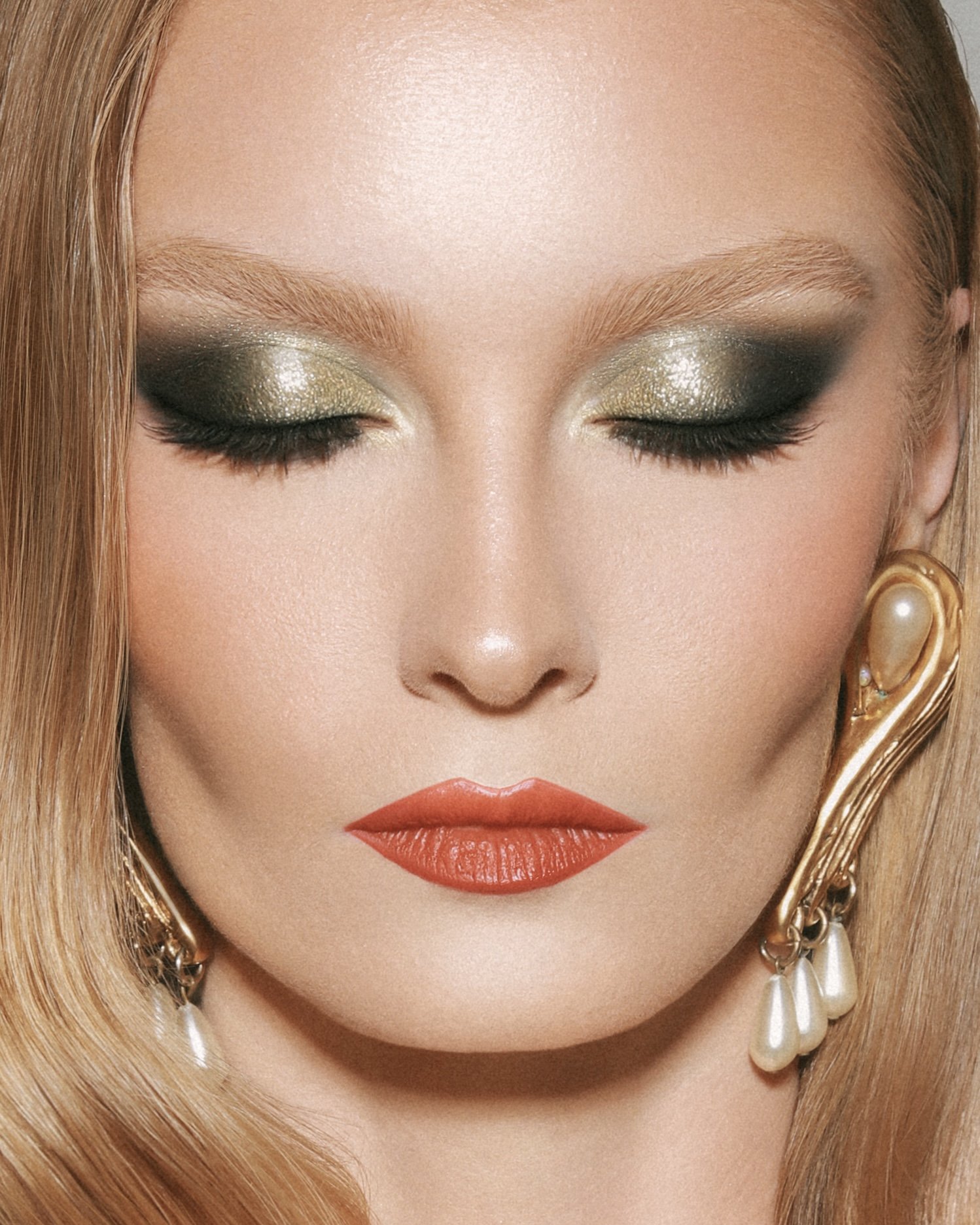 Close-up portrait of a blonde woman with closed eyes, makeup featuring olive green and gold eyeshadow, and large gold earrings.