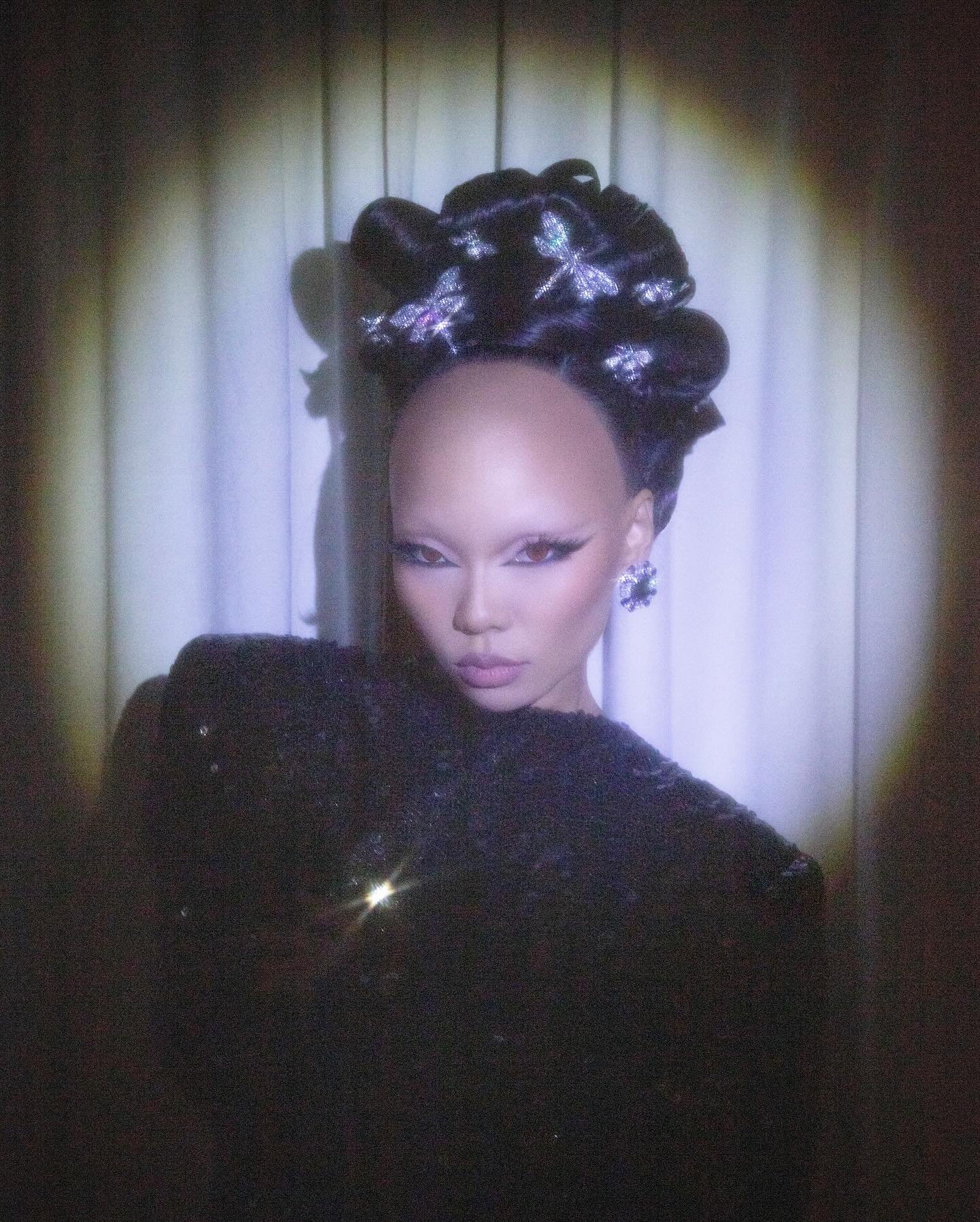 Portrait of an adult female wearing a black sequin dress and an embellished updo hairstyle, posing in front of a curtain backdrop.