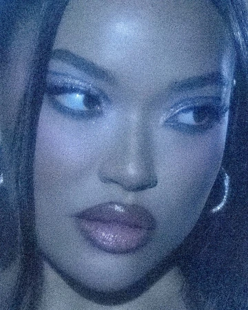 Close-up portrait of a woman with long hair, shiny makeup, and hoop earrings in a blue-toned, grainy aesthetic.