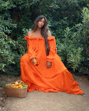 Woman in an off-the-shoulder vibrant orange tiered dress seated near a woven basket filled with fresh oranges in a garden setting.
