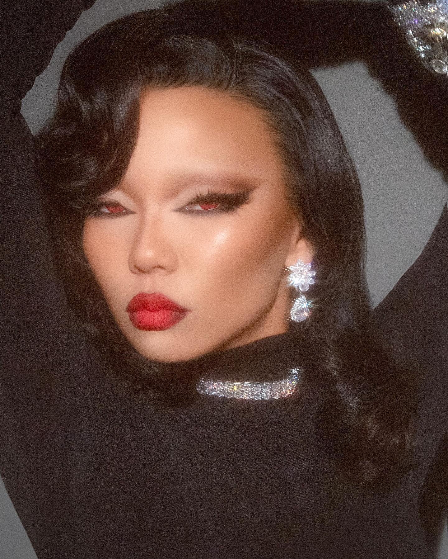 Close-up portrait of a woman with red lipstick, dramatic eye makeup, and large crystal earrings and necklace against a dark backdrop.