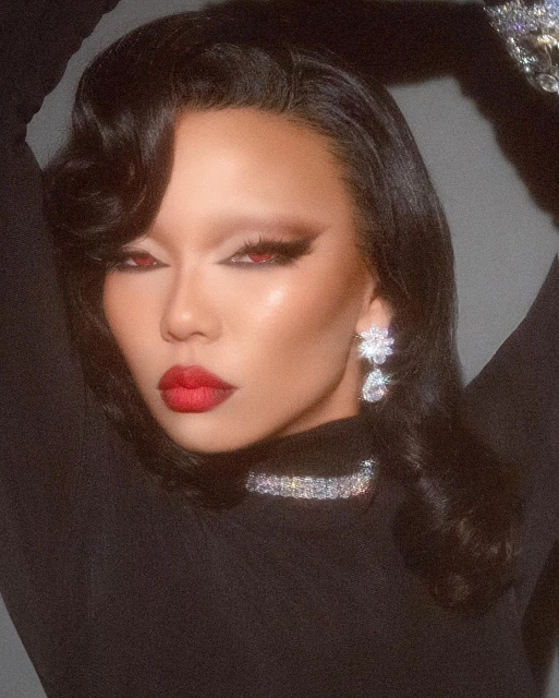 Close-up portrait of a woman with red lipstick, dramatic eye makeup, and large crystal earrings and necklace against a dark backdrop.
