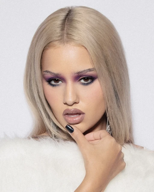 Close-up portrait of a blonde woman with purple eyeshadow and glossy lips, posing with her hand near her chin in a photo studio.