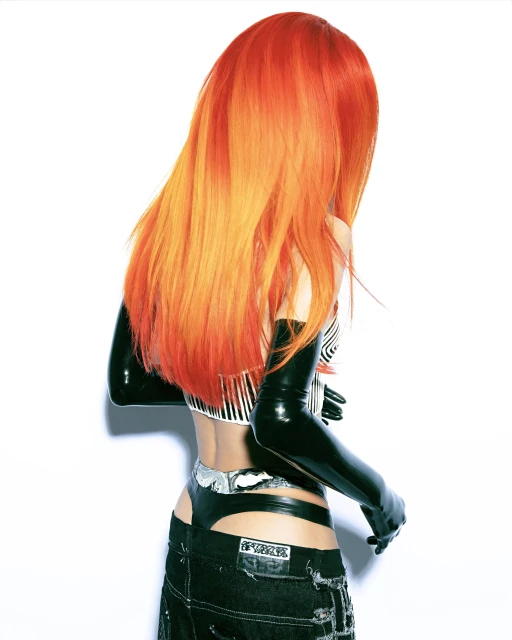 A young adult woman with orange hair and open-back jeans poses in a studio with bright lighting and a white background, rear view.