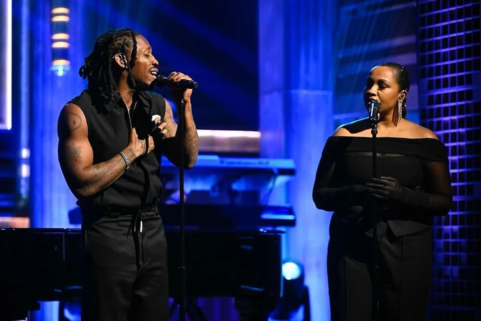 Two musicians, a man and a woman, sing into microphones on a stage with musical instruments. Stage lighting and modern set design.