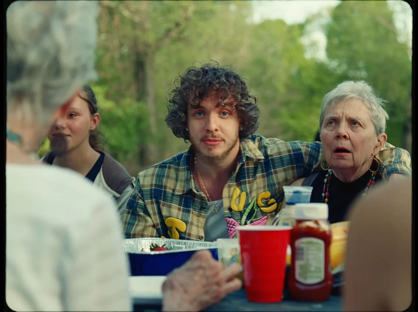 A group of people, including Jack Harlow, are gathered outdoors at a picnic table with food and drinks on a sunny day.