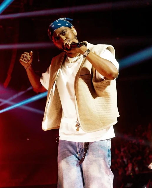 Man sings on stage in a light-colored vest, white t-shirt, blue jeans, and blue bandana. Bright performance lighting.