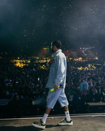 Musician walks on stage in front of a huge concert crowd, wearing denim jacket, denim shorts, and green sneakers, lit by stage lights.