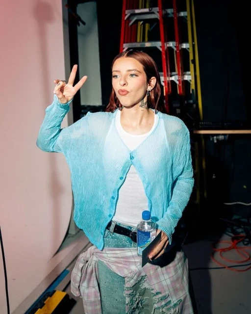 Young adult woman in a light blue cardigan, jeans, and white tank top flashing a peace sign with auburn hair and tattoos.