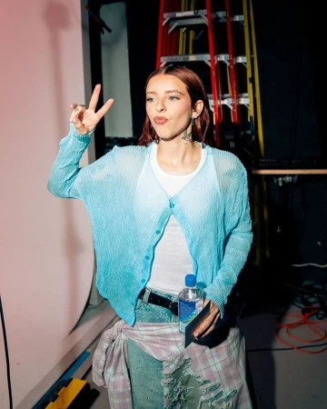 Young adult woman in a light blue cardigan, jeans, and white tank top flashing a peace sign with auburn hair and tattoos.