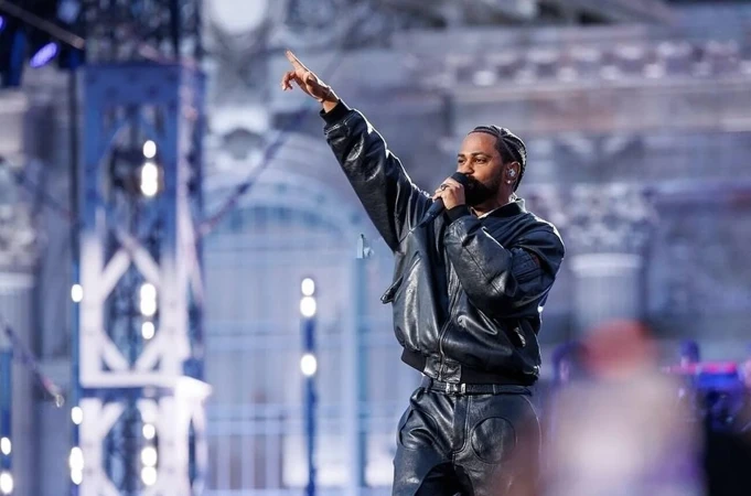 A man sings into a microphone on stage. He's wearing a black leather jacket and pants, and pointing with his left hand to the left.